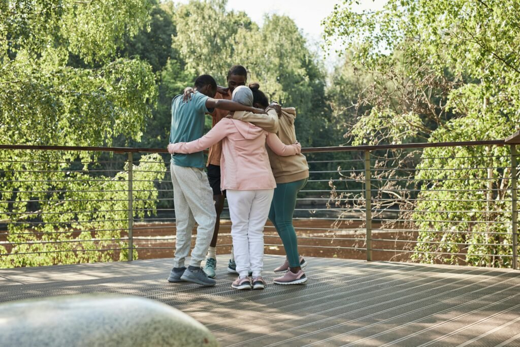 Friends huddling in a group hug on a sunny outdoor deck surrounded by greenery.